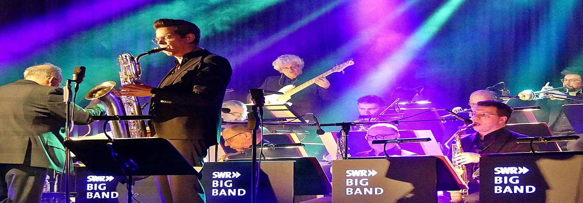 Musikband mit Instrumenten und einem Dirigenten auf einer Bühne mit buntem Hintergrundlicht