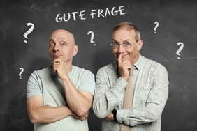 2 Männer blicken in die Kamera, mit hellem Shirt bekleidet, Arme überschlagen und eine Hand greift zur Nase, Blicken fragend in die Kamera