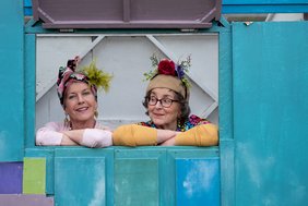 2 Damen mit bunter Kittelschürze und Kopftuch sowie Blumen schauen aus einem Fenster umrandet mit blauer Wand