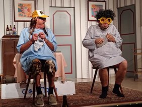 in einem altmodisch dekorierten Wohnzimmer sitzen zwei Männer, davon einer als Frau verkleidet auf einem Stuhl, sie mit übergroßer Brille undgepunktetem Nachthemd